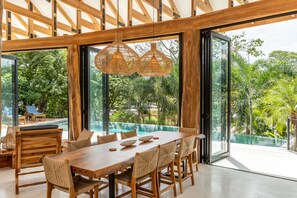 A spacious dining area with a large wooden table and chairs, large windows offering a view of a swimming pool and lush greenery. - Mediterranean Villa in Gated Guiones Community (Nosara)