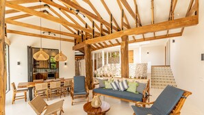 Un vaste espace de vie agrémenté de poutres en bois, d’un canapé avec des coussins à motifs, de fauteuils en osier et d’une table à manger.