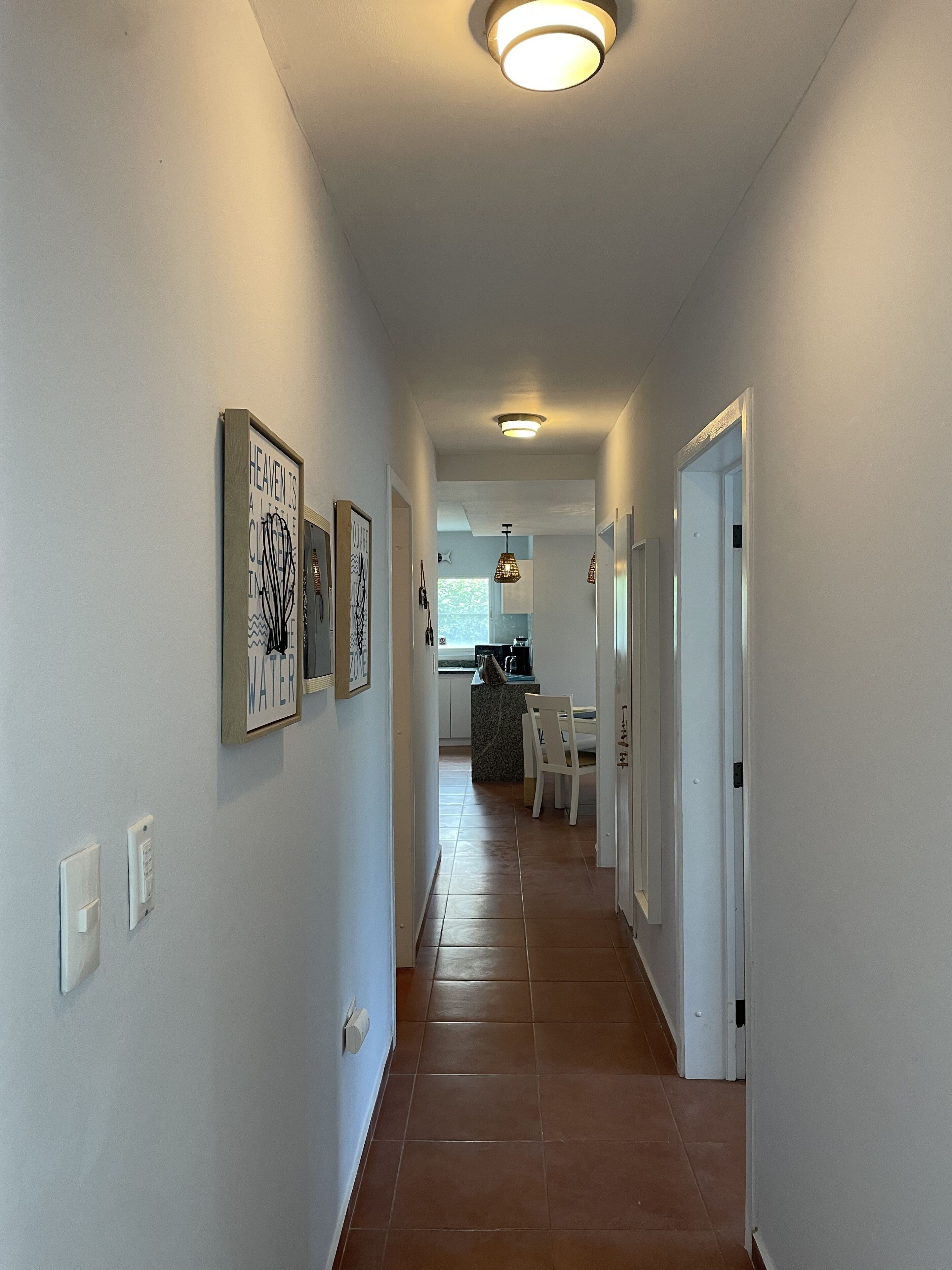 A hallway leading to a kitchen with white walls, a tiled floor, and pendant lights.