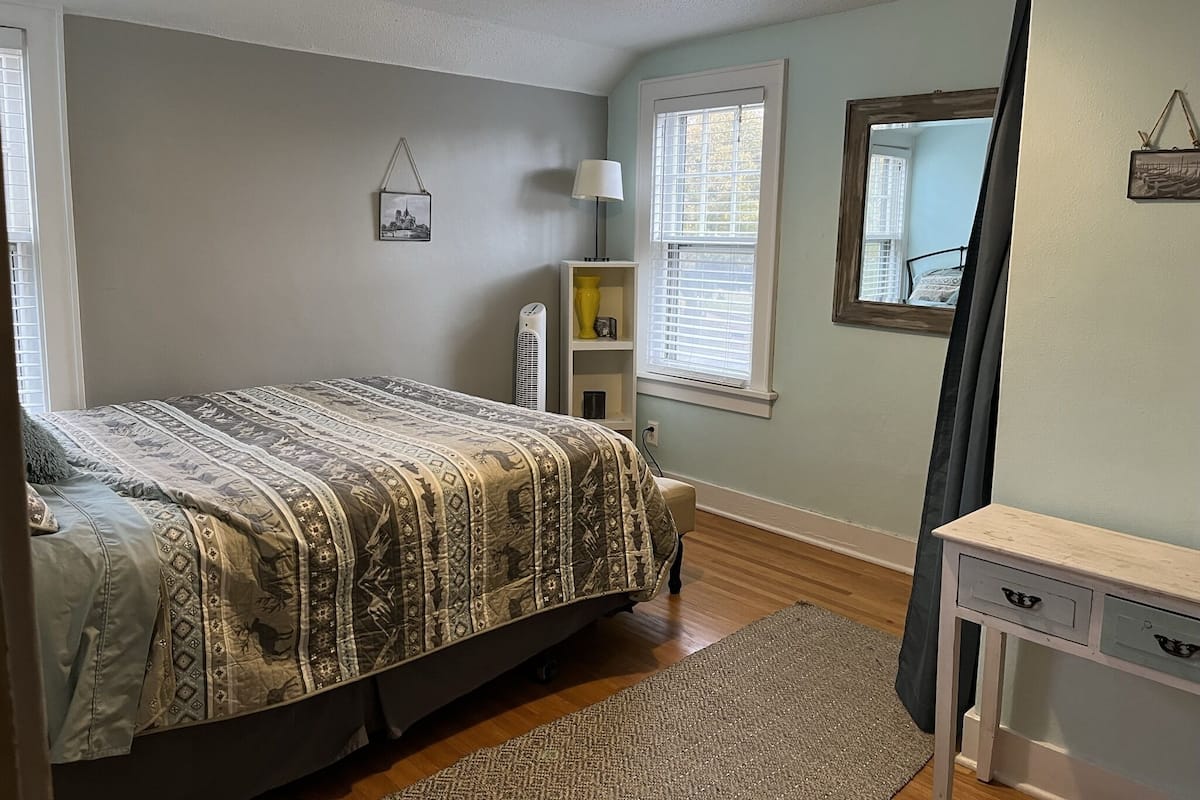 A bedroom with a bed, a bedside table, a lamp, a shelf with decorative items, and a window with blinds.