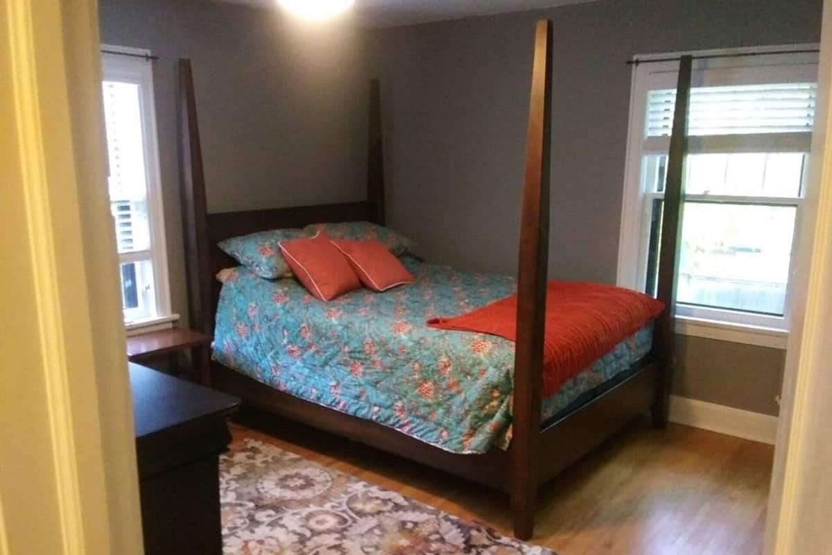 A four-poster bed with a floral bedspread and red pillows, a patterned rug, and a window with blinds.
