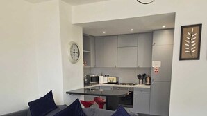 A modern living room with a grey sofa, a dining table, and a kitchen area in the background. - Two Bedroom Balcony Apartment Liverpool  (Liverpool)