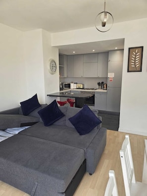 A modern living room with a grey sofa, a dining table, and a kitchen area in the background.