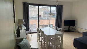 A modern living room with a dining table, chairs, a sofa, a TV, and a balcony with a view of buildings. - Two Bedroom Balcony Apartment Liverpool  (Liverpool)