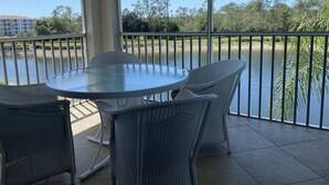 A covered outdoor area with a round glass table and wicker chairs, overlooking a body of water and a golf course. - Bright corner unit with Pond View Overlooking Hole 8 at Naples Heritage (Naples)