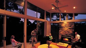 A modern outdoor living area with a stone fireplace, large windows, and a ceiling fan.