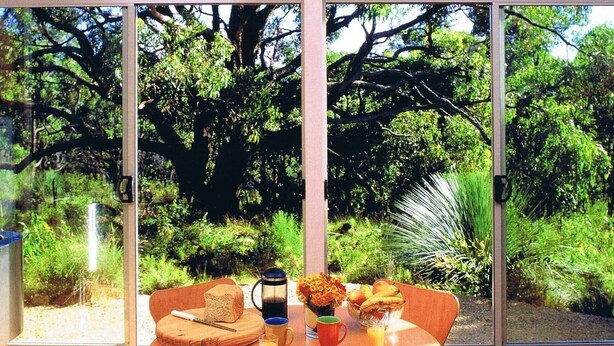 A dining table with a breakfast setting, including bread, fruit, and a coffee pot, in a room with large windows overlooking a lush garden.