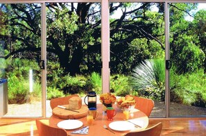 A dining table with a breakfast setting, including bread, fruit, and a coffee pot, in a room with large windows overlooking a lush garden. - Nature stay Ridgetop Retreat (Deep Creek National Park)