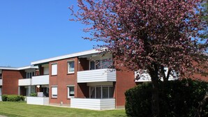 Ein zweistöckiges Backsteingebäude mit flachem Dach, einem Balkon im zweiten Stock und einem Baum mit rosafarbenen Blüten im Vordergrund.