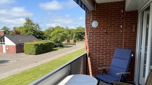 Ein Balkon mit einem weißen Tisch und zwei blauen Stühlen, einer Ziegelwand und Blick auf eine Straße sowie Grünflächen.