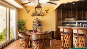 Un comedor con mesa y sillas, una lámpara colgante y una cocina con armarios de madera y salpicadero de azulejos.