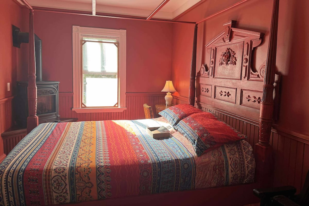 A four-poster bed with a patterned bedspread, a window with white shutters, and a wooden nightstand with a lamp.