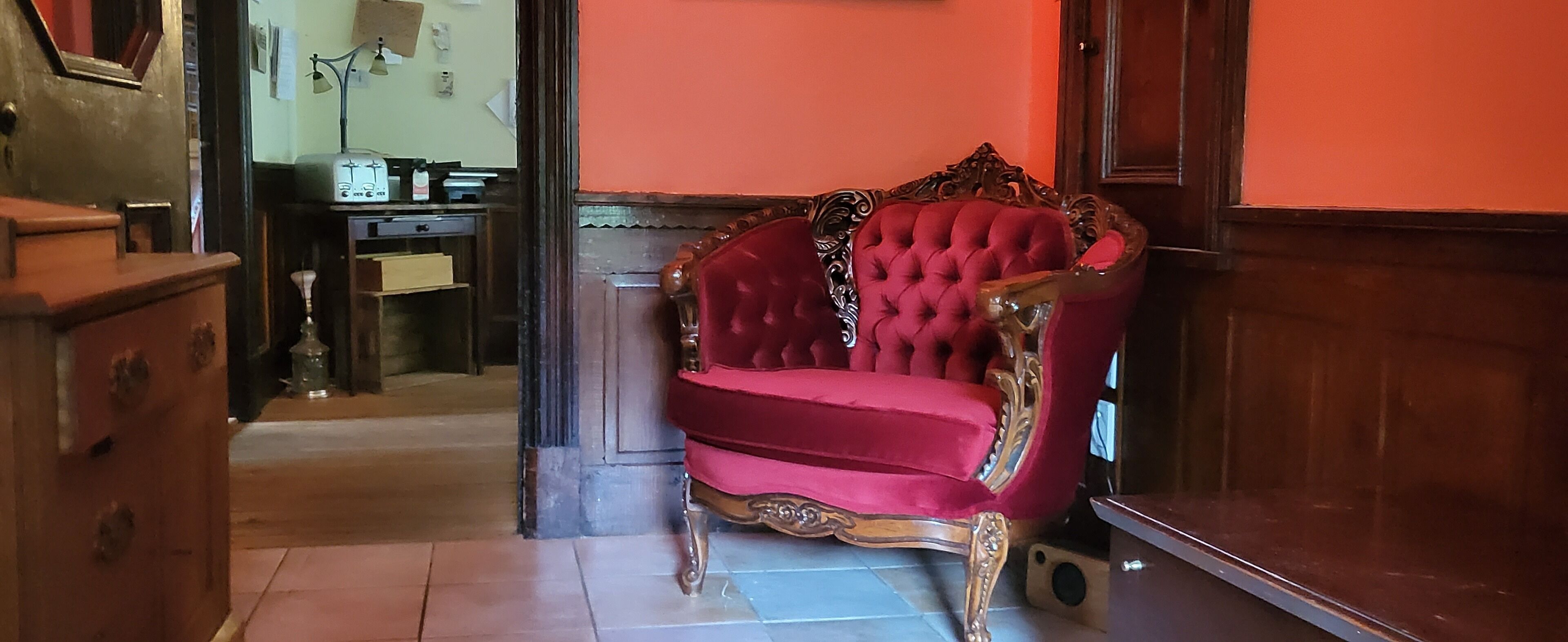 A red velvet armchair with ornate carvings in a hallway with wooden flooring and a door leading to another room.
