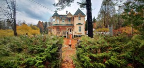 A large, multi-story house with a prominent porch and a small tower, surrounded by dense forest and autumn foliage. - Le Salon des Inconnus (Namur)