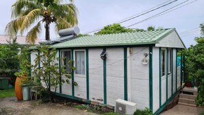 A small, single-story house with a green roof, white walls, and green trim. It has a small porch with a bench, a solar panel, and a satellite dish. - Gite Creole (VIEUX-HABITANTS)
