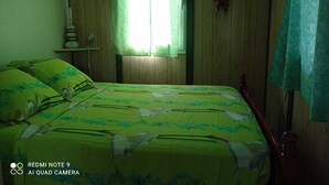A room with a bed, a window with curtains, a wall-mounted air conditioner, and a small shelf with decorative items. - Gite Creole (VIEUX-HABITANTS)