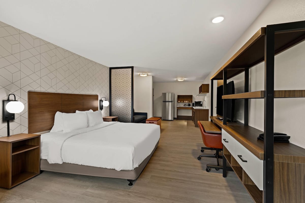 A hotel room with a bed, bedside table, lamp, desk, chair, and kitchenette.