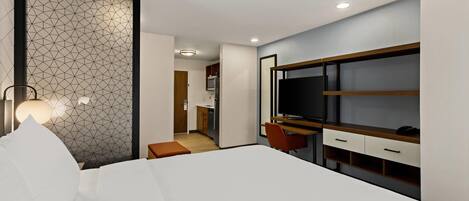 Una habitación de hotel moderna con una cama grande, un escritorio con televisor y una kitchenette al fondo.