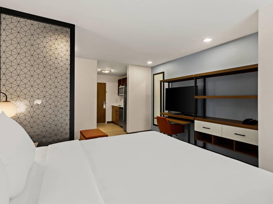 Een moderne hotelkamer met een groot bed, een bureau met een televisie en een kitchenette op de achtergrond.