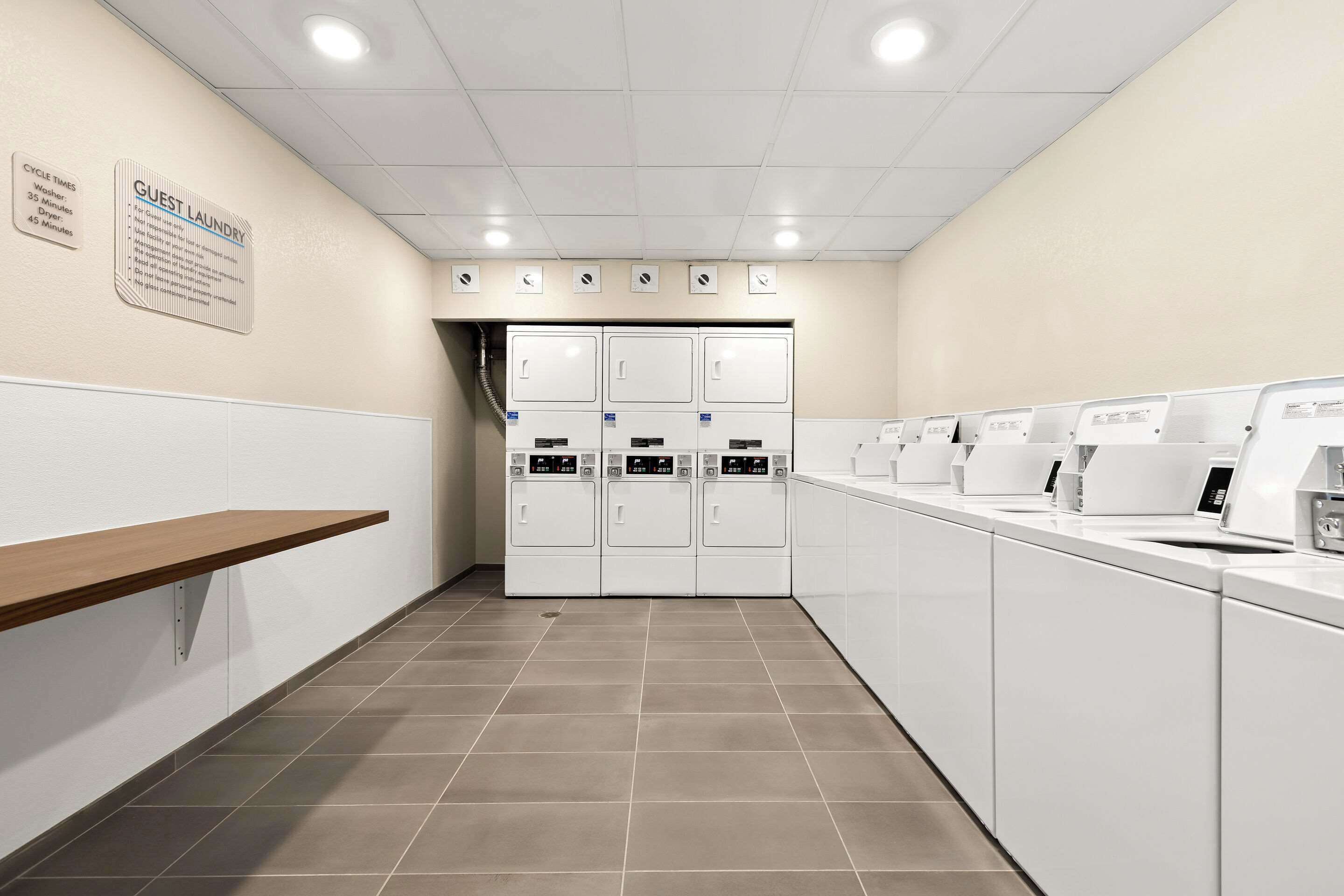A hotel laundry room with a row of white washers and dryers, a wooden shelf, and a wall-mounted sign detailing cycle times.