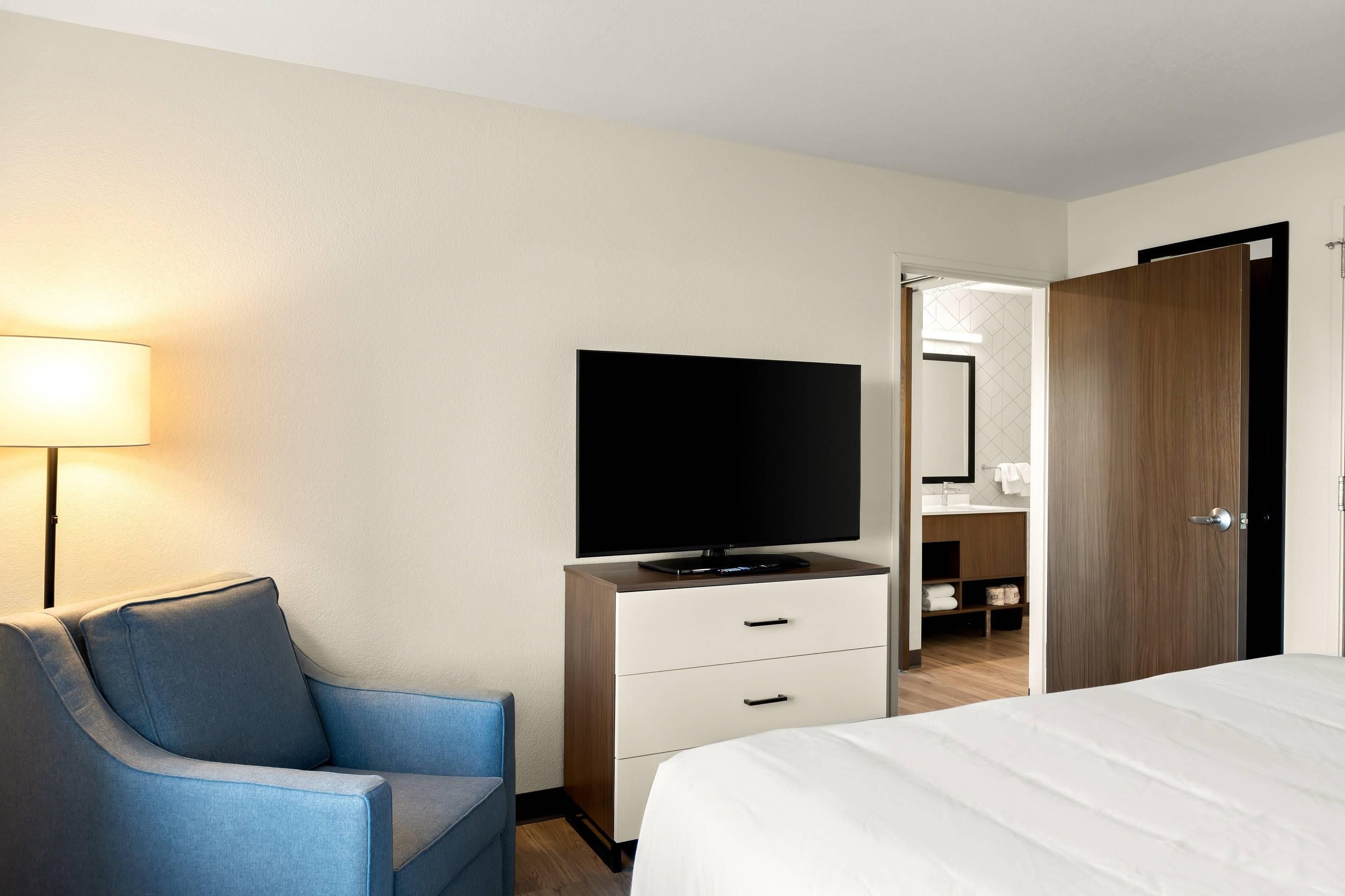 Een hotelkamer met een bed, een tv op een standaard, een bureau met lades, een stoel, een lamp en een zichtbare badkamer door een open deur.