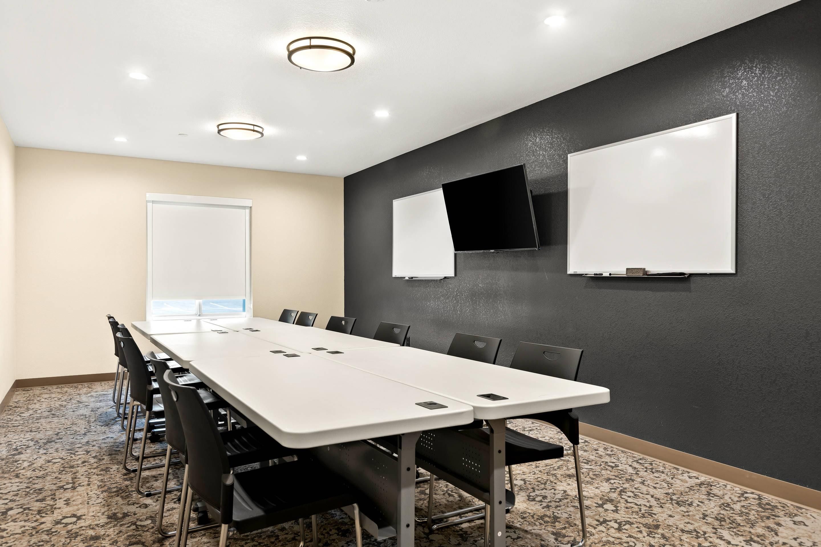 A conference room with a long table, chairs, a whiteboard, a flat-screen TV, and a window.