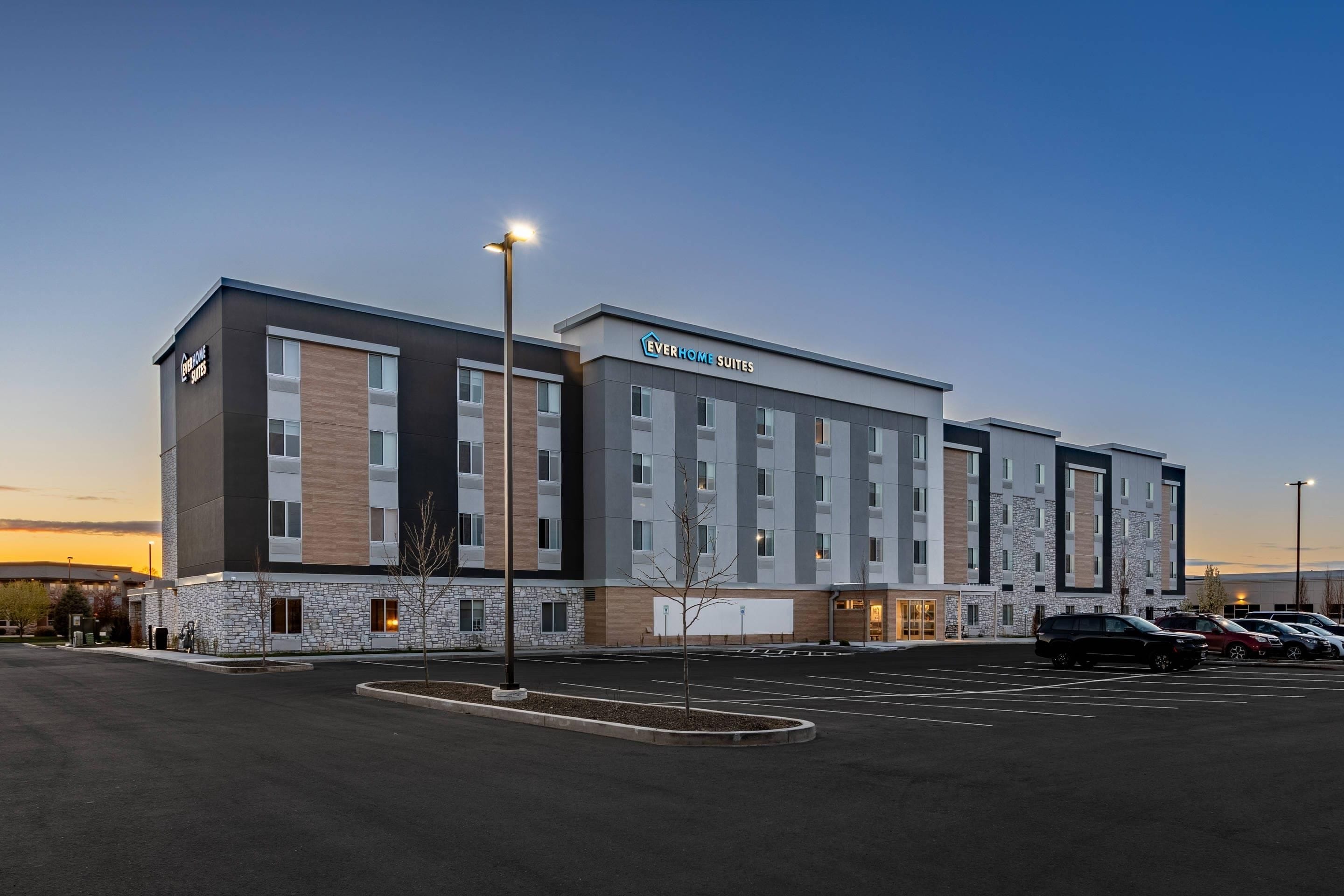 A modern hotel building with multiple floors, a parking lot, and a clear sky.