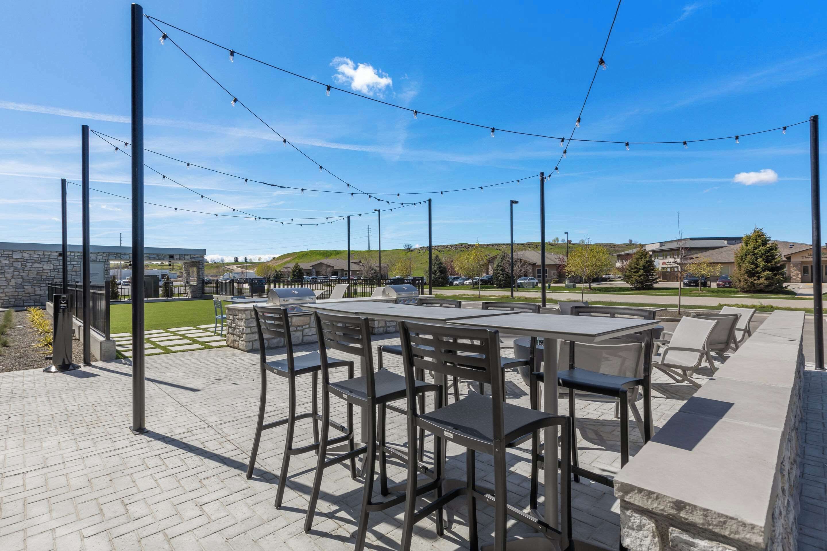 En udendørs terrasse med borde og stole, lyskæder og en stenvæg.