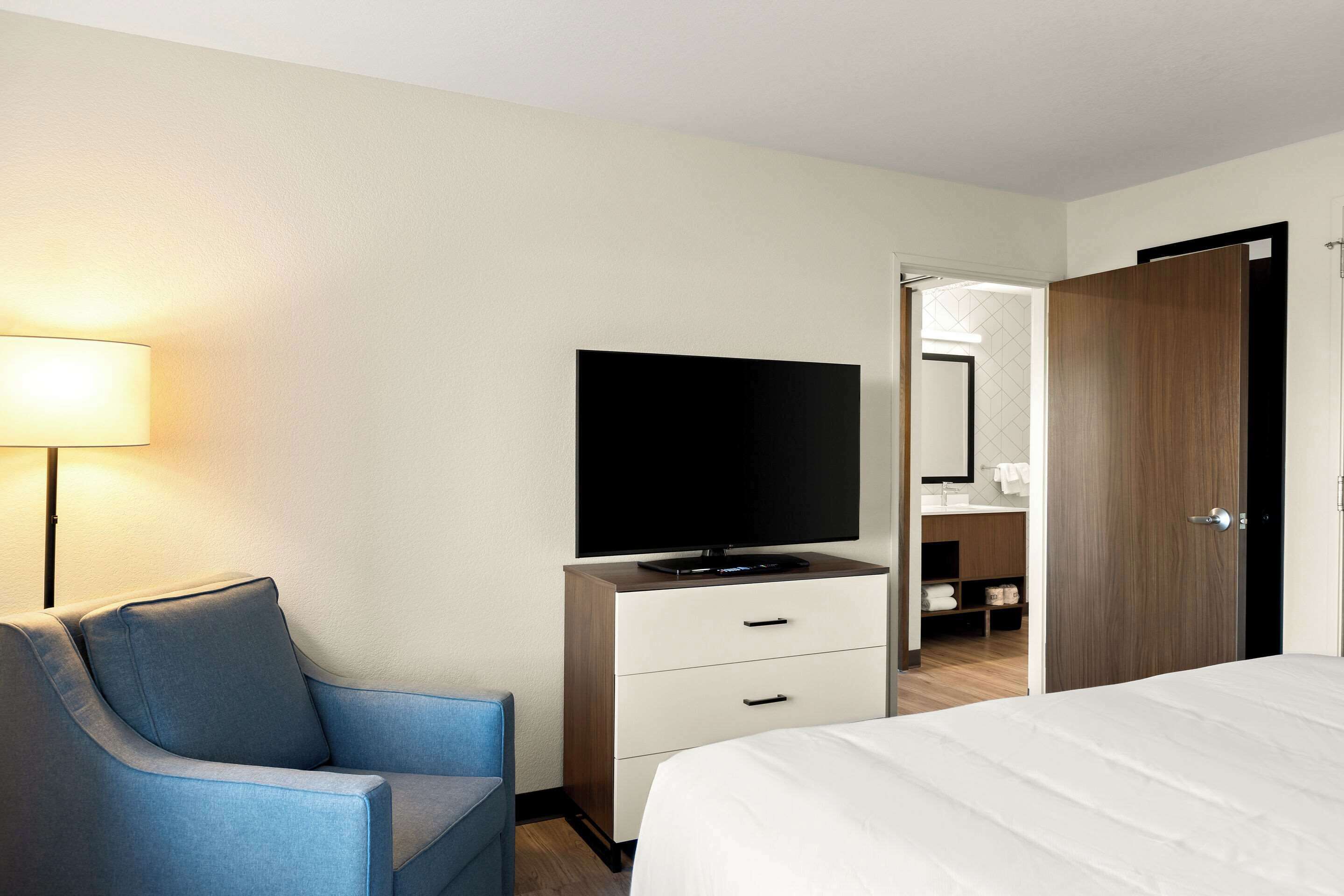 A hotel room with a bed, a TV on a wooden dresser, a blue armchair, and a bathroom visible through an open door.