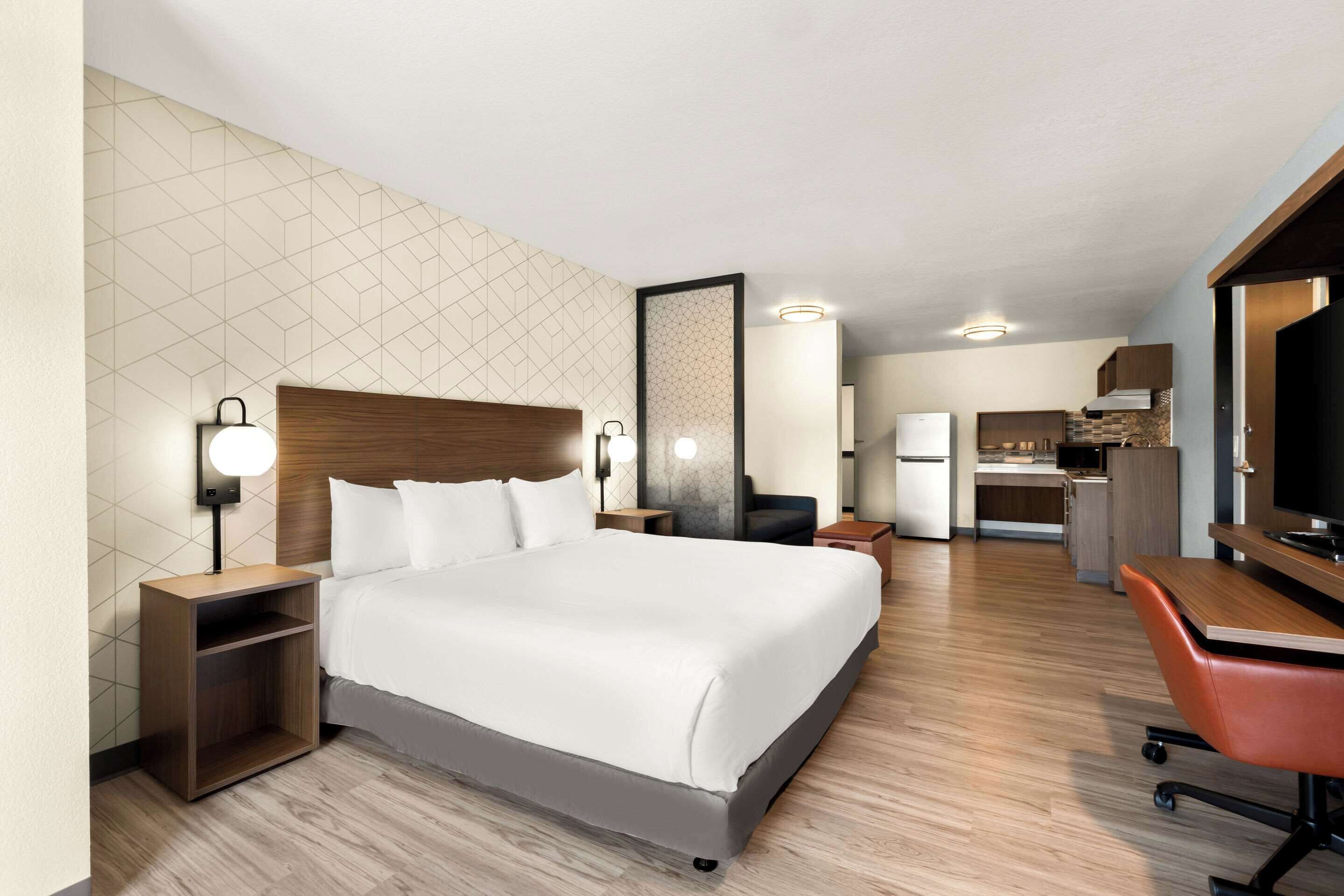 A modern hotel room with a large bed, bedside tables, a desk, and a kitchenette.