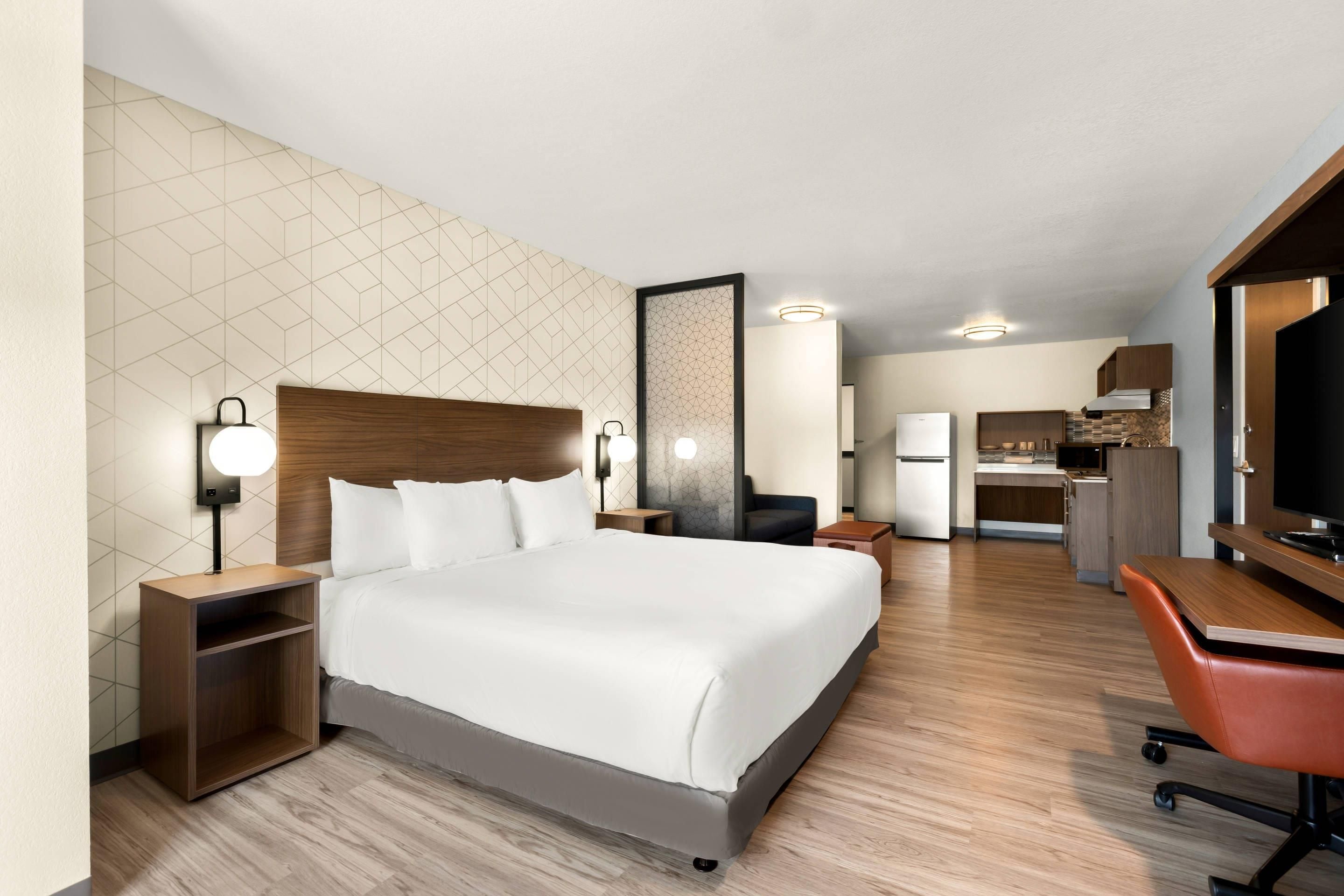 A hotel room with a large bed, bedside tables, a desk, a chair, and a kitchenette.