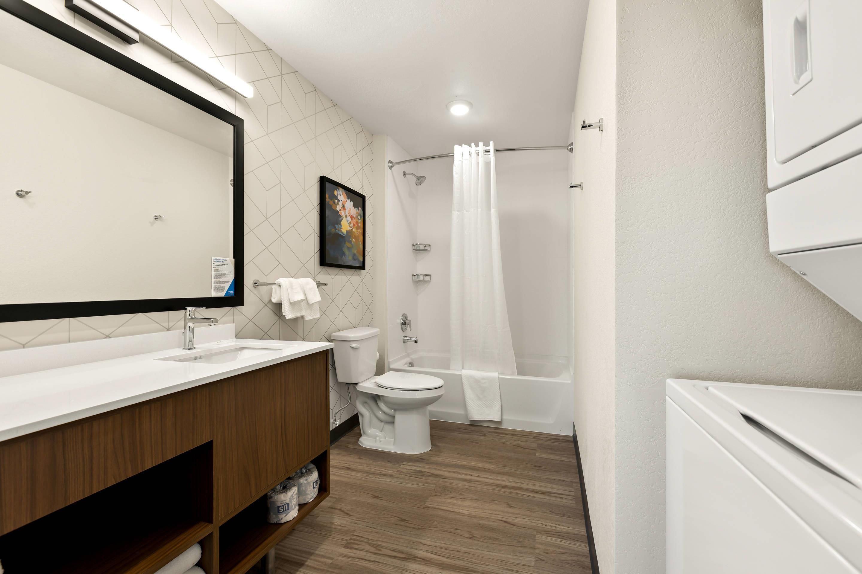 Une salle de bain moderne avec un grand miroir, des accessoires blancs, une douche avec un rideau et un meuble-lavabo en bois.