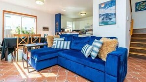 Un salon avec un canapé d’angle bleu, un coin repas avec une table et des chaises, une cuisine à l’arrière-plan et un escalier.