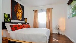 Une chambre à coucher avec un lit, un grand tableau représentant Bouddha, une fenêtre avec des rideaux et une décoration florale.