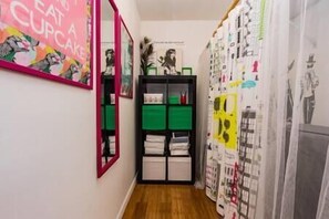 A narrow hallway with a mirror on the left, a black shelving unit with green and white storage boxes, and a wall with various posters and artwork.