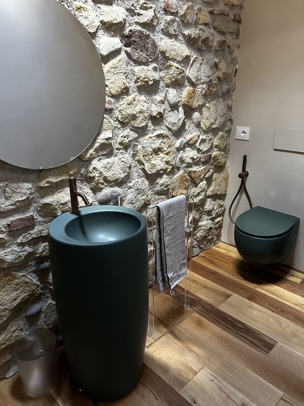 Un baño con pared de piedra, un lavamanos redondo, un perchero para toallas y un inodoro.