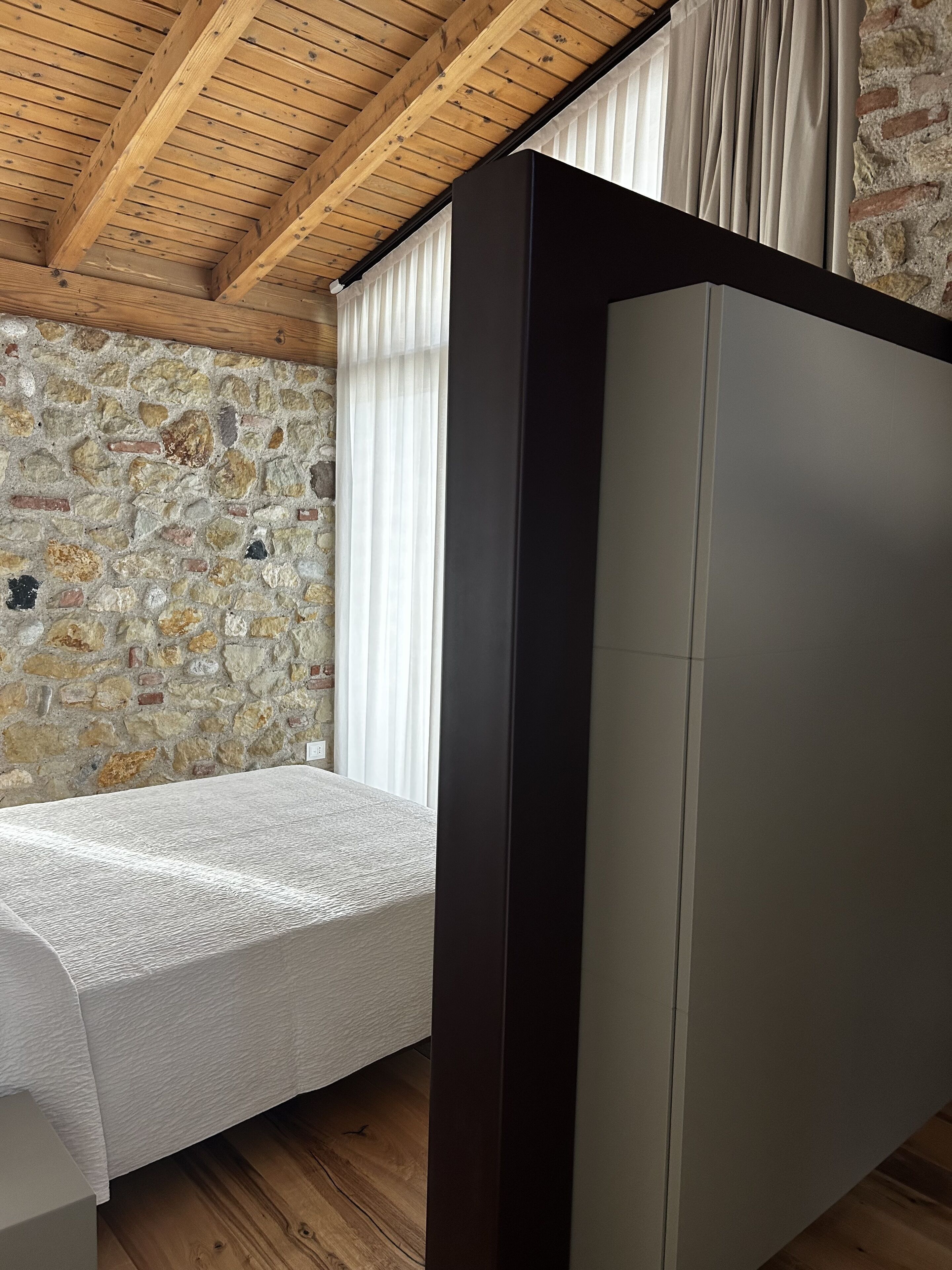 A bedroom with a stone wall, wooden ceiling beams, a bed, and a large wardrobe.