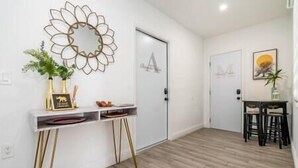 Un interior moderno con suelo de madera, una consola blanca con patas doradas, un espejo decorativo y un cuadro enmarcado en la pared.
