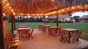 An outdoor dining area with wooden tables and chairs under a thatched roof, surrounded by a grassy lawn and palm trees. - PUNTA COCO VILLAS BEACH (Cojimies)