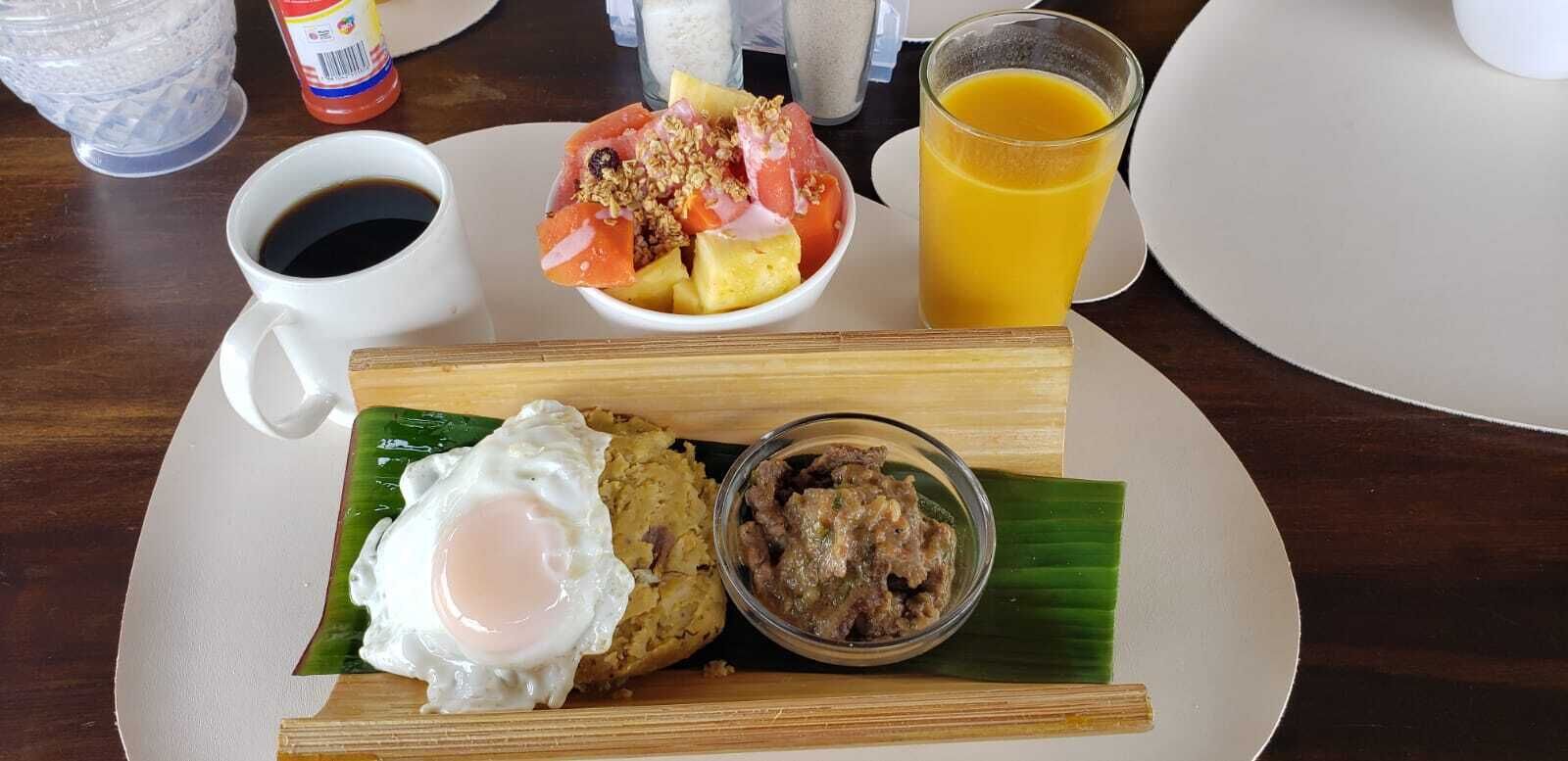 Une assiette de petit-déjeuner comprenant un œuf au plat, un bol de salade de fruits, un petit bol de viande, un verre de jus d’orange et une tasse de café.