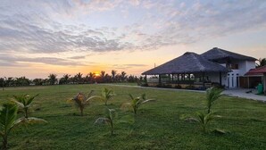 A grassy area with a thatched-roof structure, a building with a red roof, and a sunset in the background. - PUNTA COCO VILLAS BEACH (Cojimies)