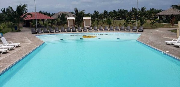 Una piscina rectangular con superficie de agua azul transparente, rodeada de sillas de descanso y un pequeño edificio con techo rojo.