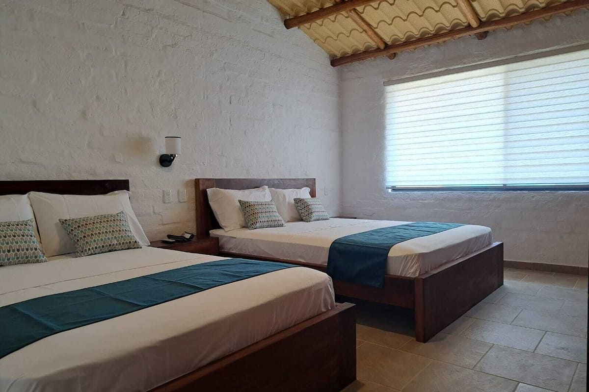 Habitación de hotel con dos camas, un ventanal con persianas y una pared blanca con textura.