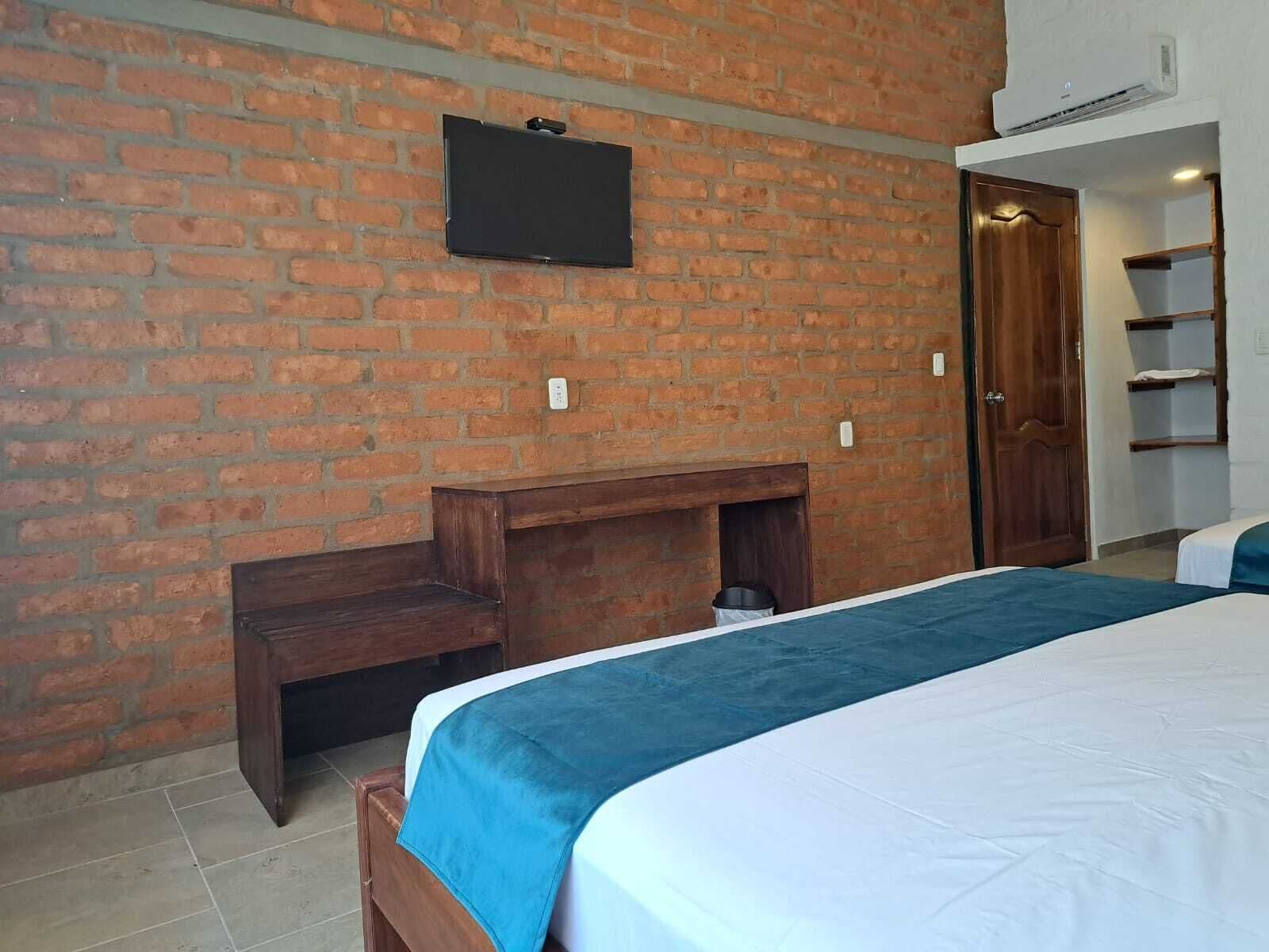 A bedroom with a brick wall, a flat-screen TV mounted above a wooden bench, a bed with a blue and white bedspread, and a wooden door.