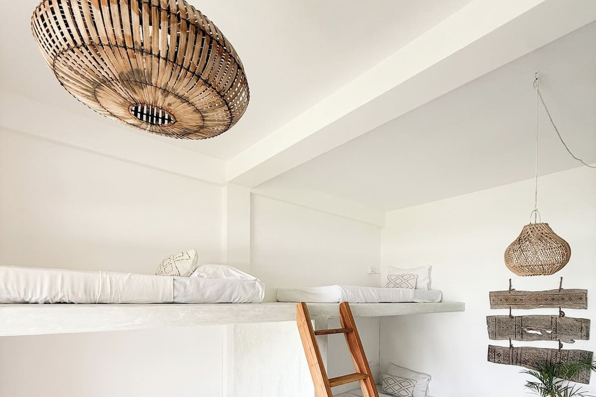 Een minimalistische kamer met stapelbedden, een houten ladder, gevlochten manden en een hanglamp.