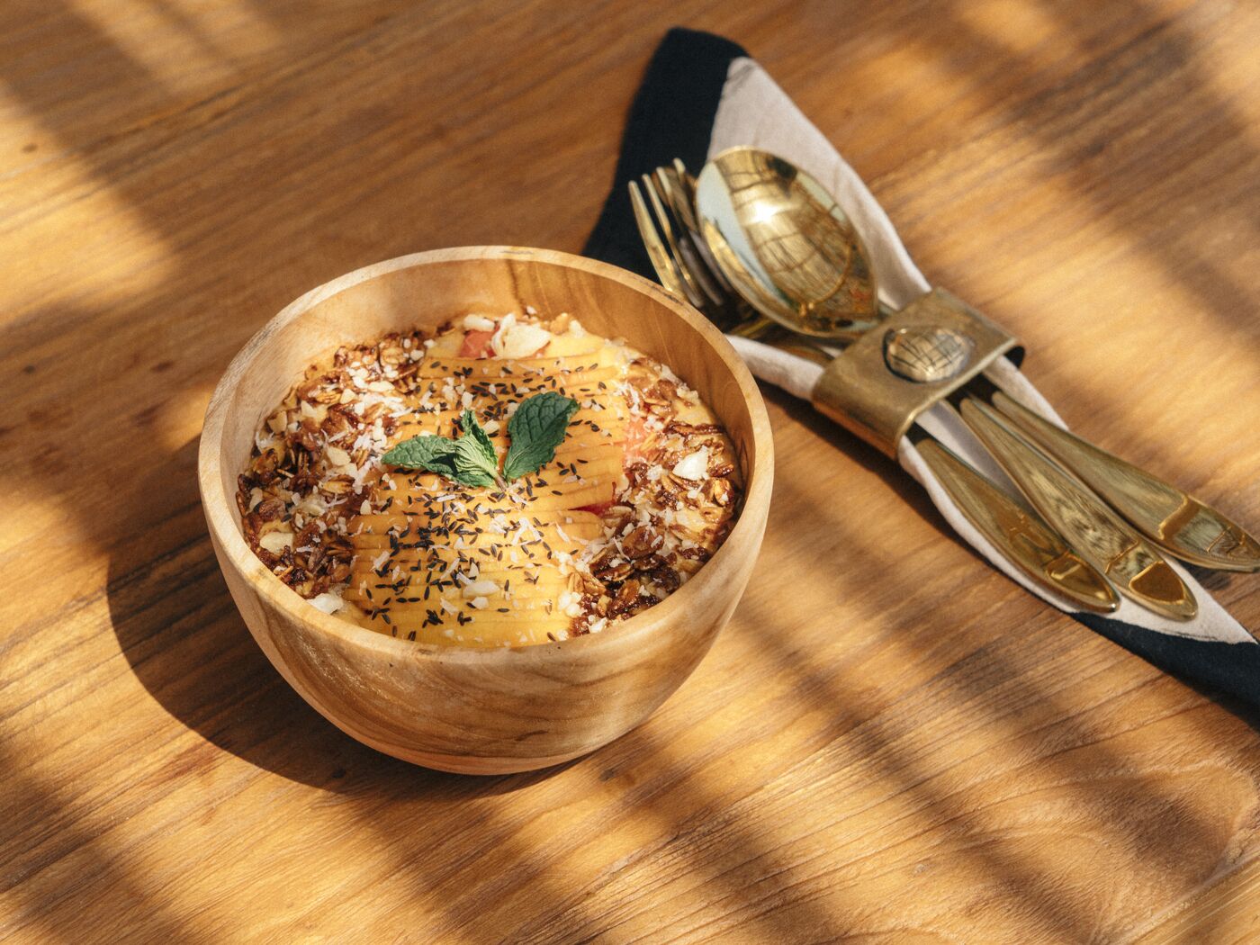 A wooden bowl with a dessert, garnished with seeds and mint, on a wooden table with a napkin and cutlery.