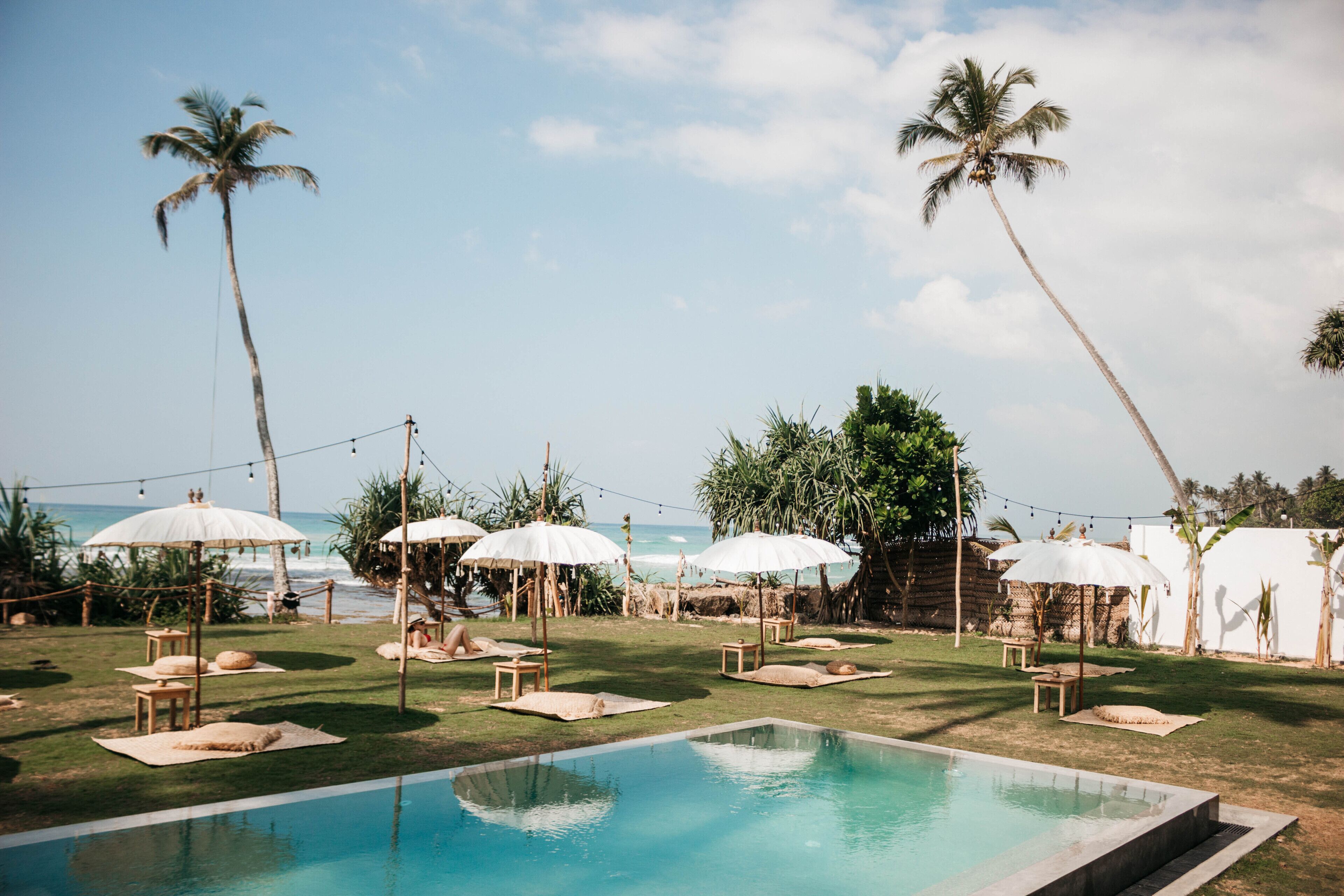 A poolside area with lounge chairs and umbrellas, palm trees, and a view of the ocean.