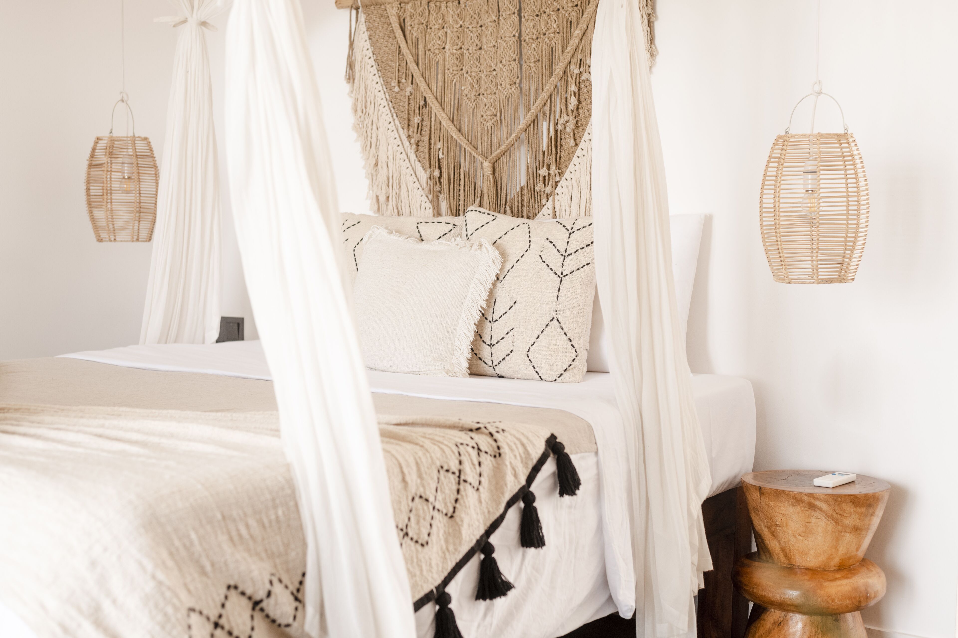 Una cama con dosel, ropa de cama blanca y almohadones decorativos. Se observan dos tapices tejidos y un taburete de madera con un libro.