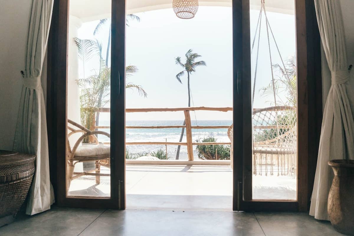 Een kamer met een grote glazen deur die leidt naar een balkon met uitzicht op de oceaan en palmbomen.