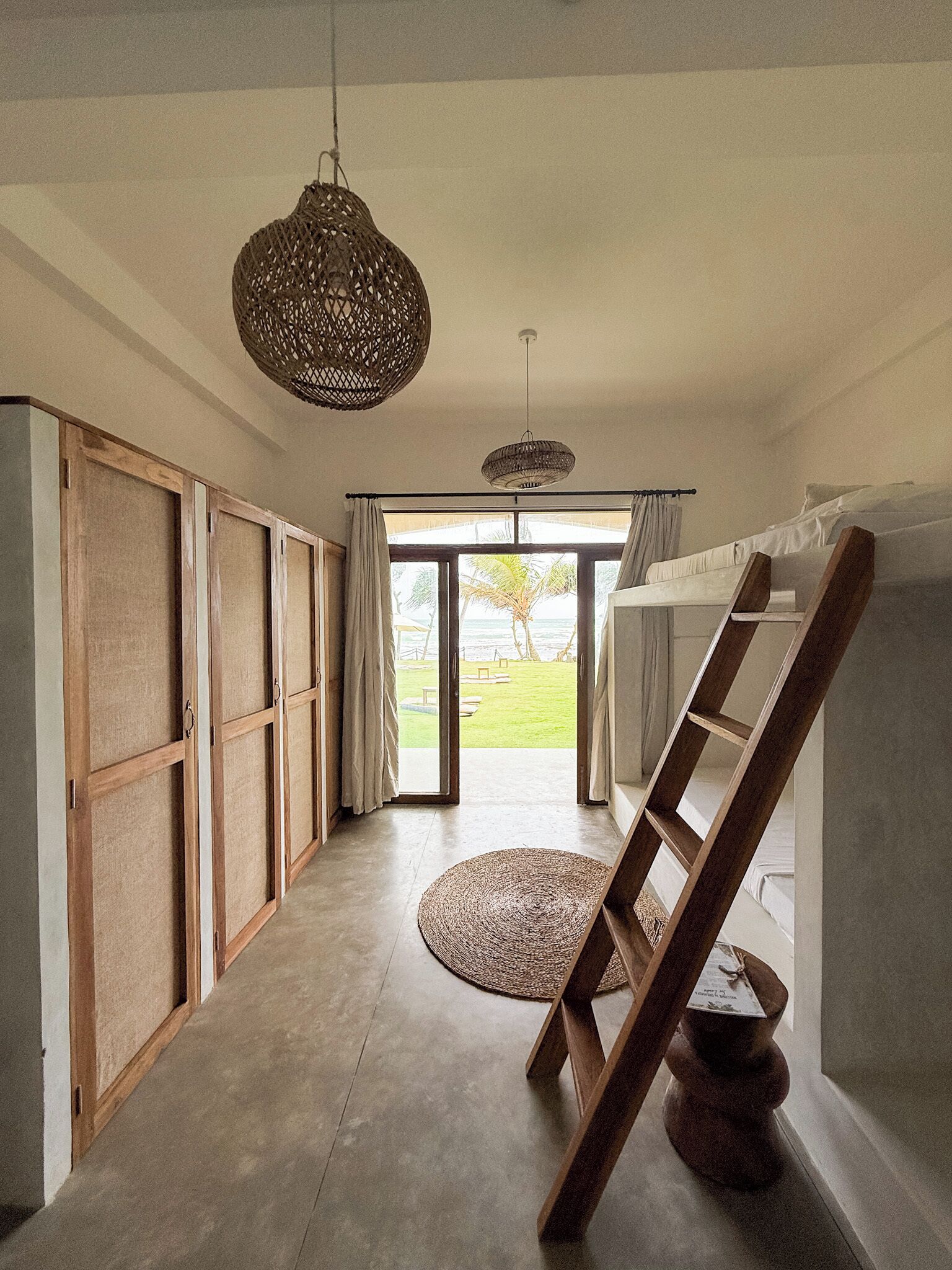 A room with a large sliding door leading to a balcony, a wooden ladder, and a woven floor mat.